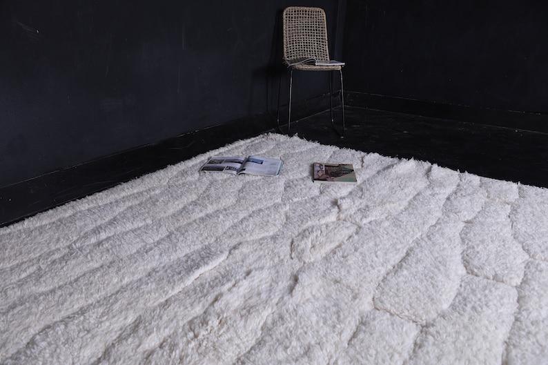 May include: A white fluffy rug with a textured pattern. The rug has a subtle wave-like design and is laid out on a dark floor.