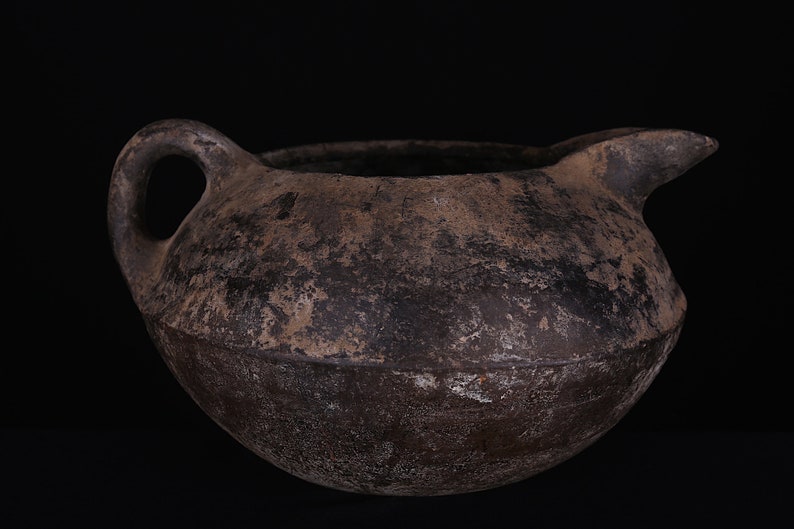 May include: A dark brown, round, ceramic pitcher with a handle and a spout. The pitcher has a rough, textured surface.
