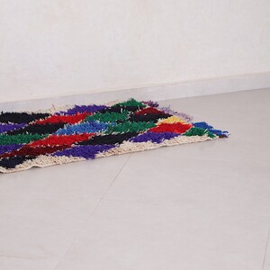 May include: A colorful, handwoven rug with a geometric pattern in shades of red, blue, green, purple, and black. The rug is made of wool and has a fringe on the ends.
