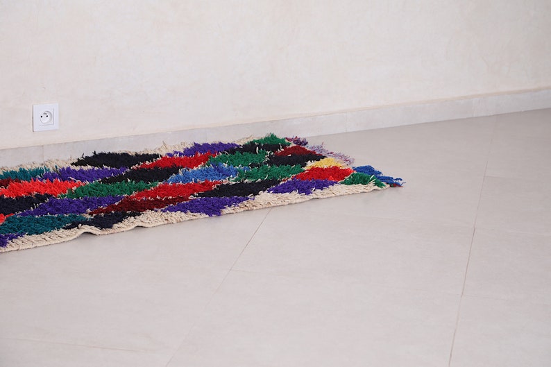 May include: A colorful, handwoven rug with a geometric pattern in shades of red, blue, green, purple, and black. The rug is made of wool and has a fringe on the ends.