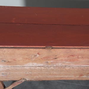 May include: An antique wooden chest with a hinged lid, painted in a deep red on top. The body of the chest is made of lighter, natural wood. The design is simple and functional, indicating its use for storage purposes.