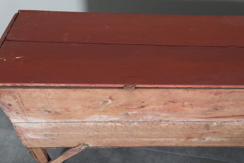 May include: An antique wooden chest with a hinged lid, painted in a deep red on top. The body of the chest is made of lighter, natural wood. The design is simple and functional, indicating its use for storage purposes.