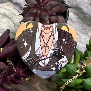Op de afbeelding: Hartvormige pin met een cartoonfiguur met wit haar, gekleed in een zwart-witte outfit met een gouden ketting. Het personage houdt een zwarte en gouden staf vast. De achtergrond is een gele en oranje gradiënt met witte sterren.
