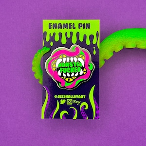 Pode incluir: Pin de esmalte com um design de boca de monstro. O pin apresenta uma boca rosa e verde com dentes brancos e olhos amarelos, com as palavras "MONSTER FUCKER" em verde. O pin está em um fundo roxo com tentáculos verdes.