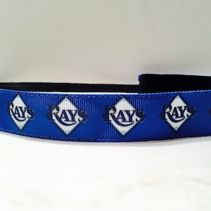Puede incluir: Diadema azul y blanca con un patrón repetido del logotipo de los Tampa Bay Rays. El logotipo es en forma de diamante con la palabra "Rays" en una fuente azul marino.