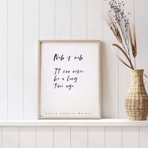 May include: A framed print featuring the quote "Now is now. It can never be a long time ago." by Laura Ingalls Wilder. The print is in a light wood frame and is displayed on a white shelf. A woven vase with dried plants is to the right.