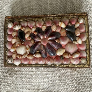 May include: A small rectangular box with a decorative design made of seashells. The box is covered in a variety of seashells, including pink, white, and purple shells. The box has a gold trim around the edges.