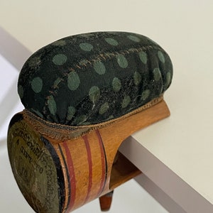 May include: A vintage green and brown pincushion with white polka dots. The pincushion is attached to a wooden base with red and white stripes and a label that reads "A GIFT TO" in cursive.