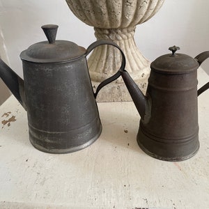 May include: Two vintage metal teapots, one gray and one brown, with a worn finish, sitting on a white surface.