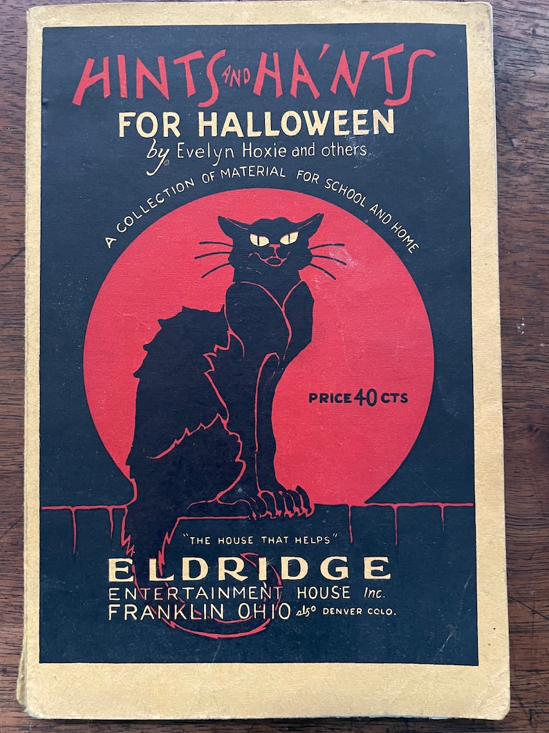 May include: A vintage book cover with a black cat silhouette against a red moon. The title is "Hints and Ha'nts for Halloween" by Evelyn Hoxie and others. The book is a collection of material for school and home. The price is 40 cents. The publisher is Eldridge Entertainment House Inc. in Franklin, Ohio, also Denver, Colorado.