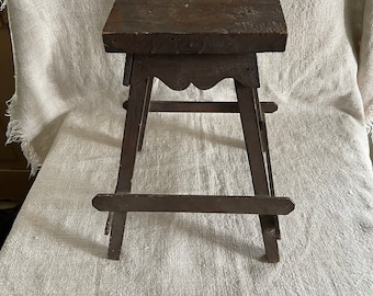 May include: A dark brown wooden stool with a square top and a simple, rustic design. The stool has four legs that are connected by a crossbar.