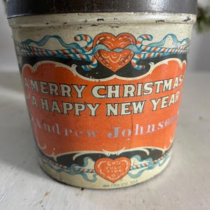 May include: A vintage tin container with a red, white, and blue Christmas design. The tin has a heart-shaped design with the words "A Merry Christmas A Happy New Year" and "Andrew Johnson" printed on it. The bottom of the tin has the words "Cou Yule Tide" and "Am Can Co. Zea" printed on it.