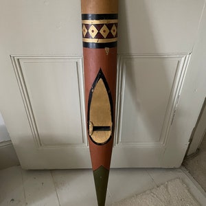 May include: A long, wooden, hand-painted, decorative object with a pointed tip. The object is painted in shades of brown, red, green, black, and gold. It has a diamond pattern and a teardrop-shaped cutout.