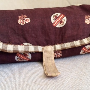 May include: A brown fabric roll-up pouch with a white and brown checkered trim and a brown leather strap. The pouch has a floral pattern with small white flowers and larger circles with red and white accents.