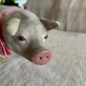 May include: A plush gray pig with a pink nose and pink ribbon around its neck. The pig is lying on a white fabric surface.