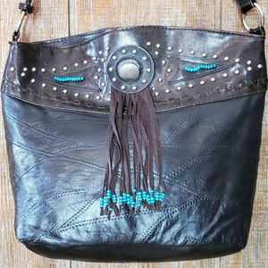 May include: A brown leather tote bag with a fringe detail and turquoise beads. The bag has a decorative metal piece with a turquoise stone in the center.