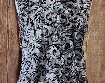 May include: A sleeveless vest with a silver and black floral pattern. The vest has a collared neckline and decorative buttons down the front. The garment is displayed on a mannequin against a wooden background.