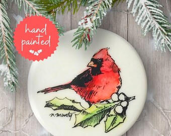 May include: Hand-painted Christmas ornament featuring a red cardinal perched on a holly branch with white berries. The ornament is white with a black outline and the artist's signature "n.murty" in the bottom left corner.