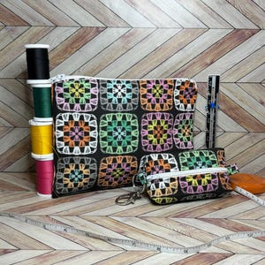 May include: Two zippered pouches with a black background and a colorful crochet pattern. The larger pouch is lying on a wooden surface with a spool of thread and a measuring tape. The smaller pouch has a key ring attached.