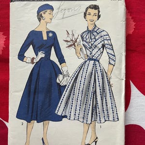 Puede incluir: Un sobre de patrón de costura Advance, talla 18, busto 36, con ilustraciones de dos vestidos. Uno es un vestido azul liso con sombrero y guantes a juego. El otro es un vestido blanco con rayas verticales azules y un cuello con lazo.