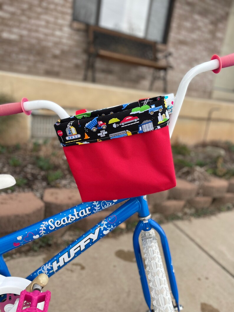 May include: A blue bicycle with a red and black handlebar basket. The basket has a black and white fabric lining with a pattern of construction vehicles. The bicycle has white tires and a white handlebar. The bicycle has the words "Seastar" and "HUFFY" printed on the frame.