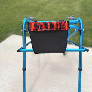 May include: A blue walker with a black storage bag. The top of the bag features a flame pattern in red and orange. The walker is on a concrete surface with a green lawn in the background.