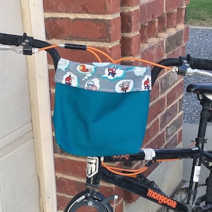 May include: A black bicycle with a teal and gray fabric bag attached to the handlebars. The bag has a print of cartoon superheroes. The bicycle has an orange handlebar grip and a black seat.
