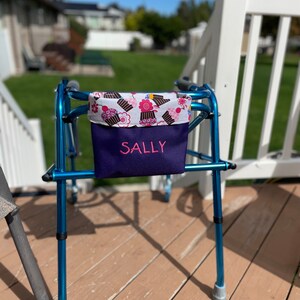 May include: A blue walker with a purple fabric bag attached. The bag has a white and pink cupcake print and the name "SALLY" embroidered on the front.
