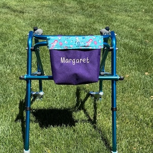 May include: A teal-colored walker with a purple bag that has the name "Margaret" embroidered on it. The bag is decorated with a teal and pink patterned fabric. The walker is on a grassy surface.