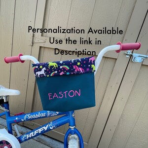 May include: A blue children's bicycle with a white basket. The basket is lined with a fabric featuring a unicorn, rainbow, and star pattern. The basket is personalized with the name "EASTON" in pink lettering.