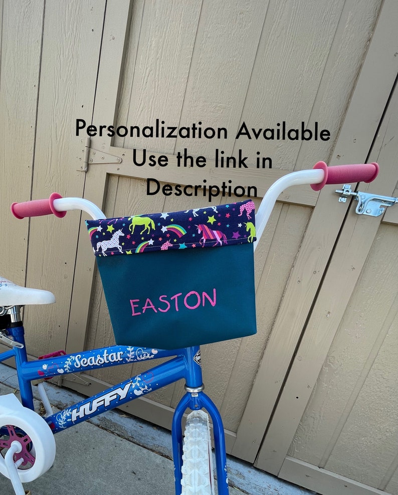 May include: A blue children's bicycle with a white basket. The basket is lined with a fabric featuring a unicorn, rainbow, and star pattern. The basket is personalized with the name "EASTON" in pink lettering.