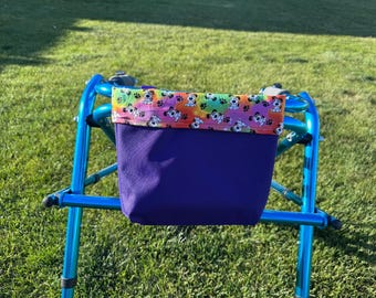 May include: A blue walker with a purple fabric bag attached. The top of the bag has a colorful border with a pattern of white puppies and paw prints. The walker is on a green grassy surface.