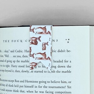 May include: A white bookmark with a repeating pattern of brown deer silhouettes. The bookmark is folded in half and placed on a book with the text "THE FOUR CH" visible. The text "INS" is also visible on the right side of the bookmark.