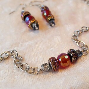 May include: A silver chain necklace with a red and gold beaded pendant. The pendant has three beads, one large red bead and two smaller gold beads with sparkly accents.