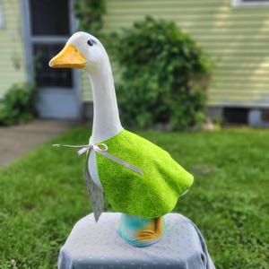 May include: A white goose lawn ornament with a yellow beak and painted eyes, wearing a bright lime green cape featuring a small, delicate floral pattern. The cape is secured at the neck with a thin, light lavender ribbon tied in a small bow.