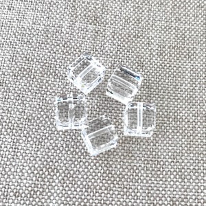 May include: Six clear plastic square beads with a hole in the center. The beads are arranged in a cluster on a light brown fabric background.