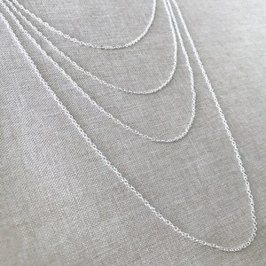 May include: A set of five silver chain necklaces of varying lengths, layered on a textured fabric background.