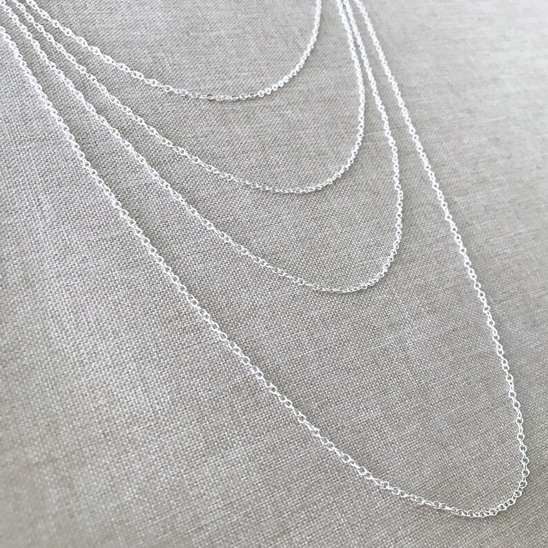 May include: A set of five silver chain necklaces of varying lengths, layered on a textured fabric background.
