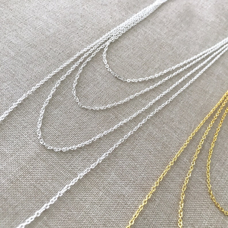 May include: A close-up of two chains, one silver and one gold, laid out on a textured fabric surface. The silver chain is layered in four rows, while the gold chain is layered in two rows.