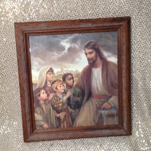 May include: A framed print depicting a religious scene. The artwork features a man in a robe surrounded by several children. The frame is made of wood with a dark brown finish. The background shows a cloudy sky and a city.