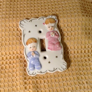 May include: White porcelain light switch cover with a raised design of two children in prayer. The girl is wearing a pink dress and the boy is wearing a blue outfit. The cover has a gold trim and stars.