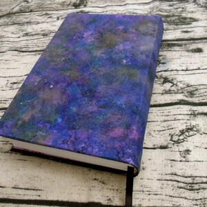 May include: A purple, blue, and green notebook with a galaxy-like design on the cover. The notebook is closed and lying on a wooden surface.
