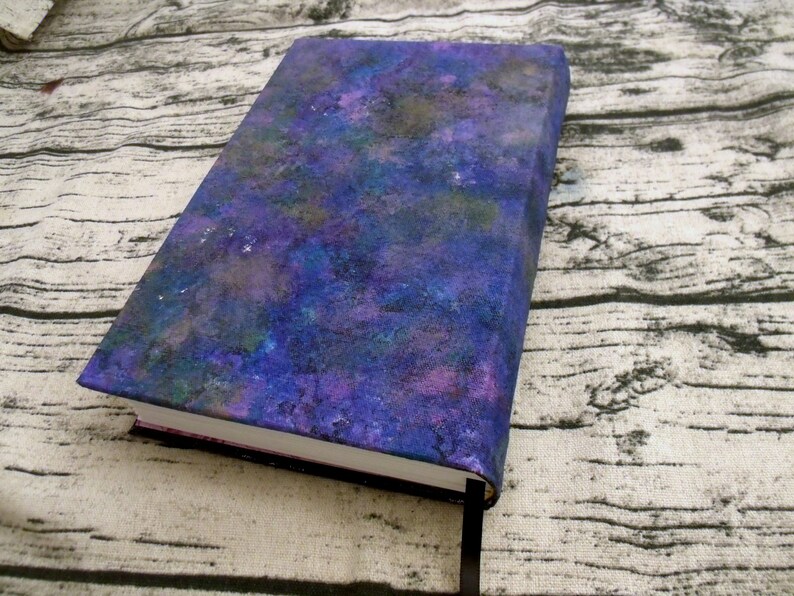 May include: A purple, blue, and green notebook with a galaxy-like design on the cover. The notebook is closed and lying on a wooden surface.