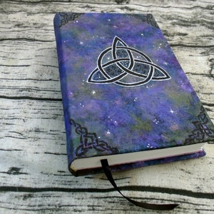 May include: A purple and blue notebook with a black Celtic knot design and a galaxy-like background. The notebook has a black ribbon bookmark.