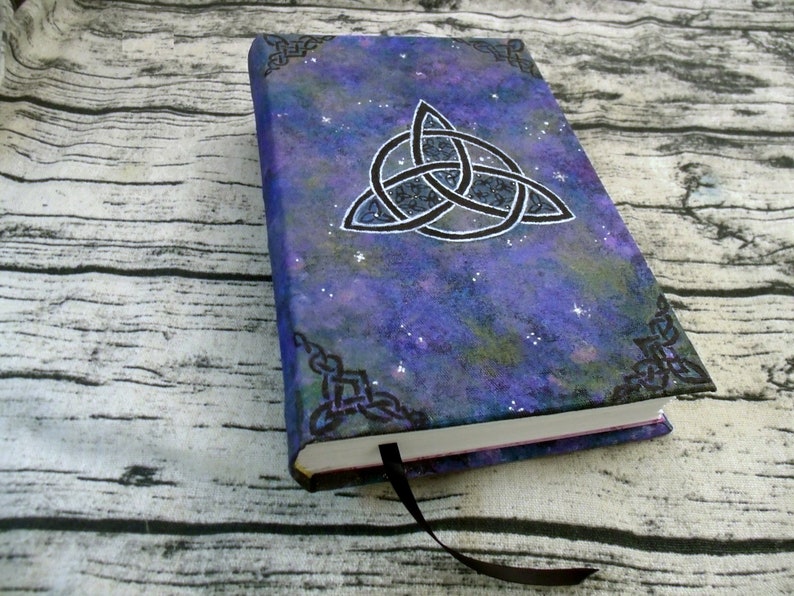 May include: A purple and blue notebook with a black Celtic knot design and a galaxy-like background. The notebook has a black ribbon bookmark.