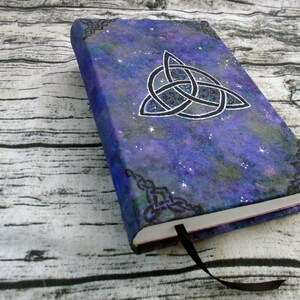 May include: A purple and blue notebook with a black and white Celtic knot design on the cover. The cover has a galaxy-like pattern with white glitter.