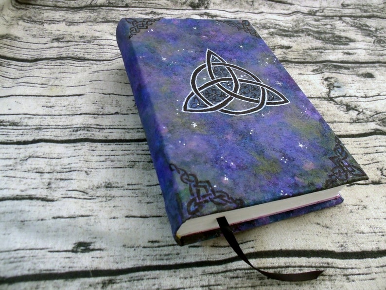 May include: A purple and blue notebook with a black and white Celtic knot design on the cover. The cover has a galaxy-like pattern with white glitter.