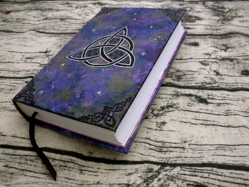 May include: A purple and black journal with a silver Celtic knot design on the cover. The cover has a galaxy-like pattern with silver glitter. The journal has a black ribbon bookmark.