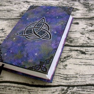 May include: A purple and black journal with a silver Celtic knot design on the cover. The cover has a galaxy-like pattern with white stars.