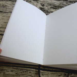May include: A blank, white, hardcover journal with a black ribbon bookmark. The journal is open to a fresh, blank page.